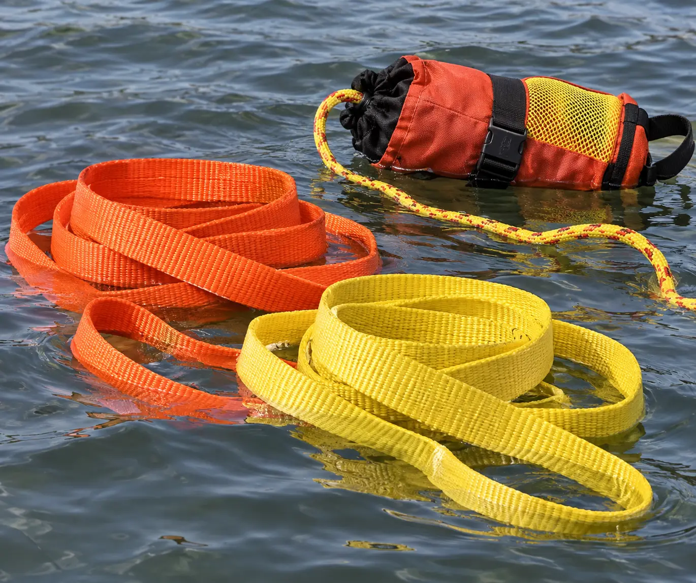 Cintas flotantes de polipropileno en naranja y amarillo de alta visibilidad enrolladas sobre la superficie del agua junto a una línea de rescate con bolsa de lanzamiento, mostrando cinta flotante utilizada en equipos de rescate acuático.
